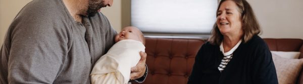 Man sitting on a couch holding a baby doll wrapped in a blanket, woman sitting nearby watching