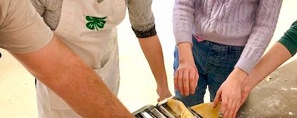 man and three youth standing around a table topped with a pasta roller, dough, and a wooden drying rack. They are rolling the dough to make pasta.