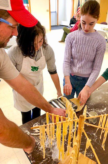 man and three youth standing around a table topped with a pasta roller, dough, and a wooden drying rack. They are rolling the dough to make pasta.
