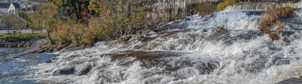 sunny outdoor scene of trees and buildings directly above a waterfall cascading over levels of rock and ledge