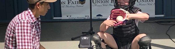 Child sitting on steps wearing baseball catcher's gear, ball in mitt, talking with young man with microphone at Union Fair