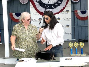 Older woman holding microphone for young man in dark glasses, rabbit on the table in front, trophies and bandstand in the background at Union Fair