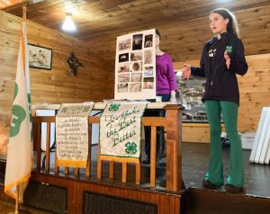 Young person speaking from a stage with 4-H flags and a photo poster.