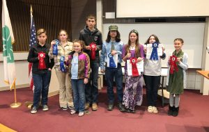 Eight young people standing in front of a stage holding award ribbons