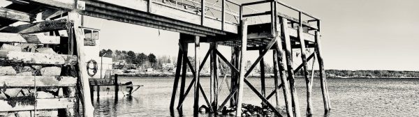 black and white photo of a deep-water dock in a harbor with land, houses in the distance