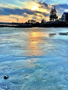 sun setting through dramatic clouds behind a point of land with a house and trees, a stretch of wet sand with footprints in the forground