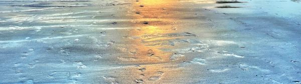 sun setting through dramatic clouds behind a point of land with a house and trees, a stretch of wet sand with footprints in the forground