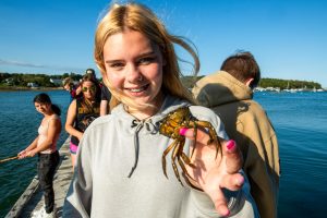 several young people on a long dock in an ocean cove, girl in foreground holding a small crab, girl to the left holding a fishing pole