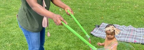 Woman and child standing outdoors on a lawn, woman is standing near a large bucket and holding two sticks with a looped strap, making a large soap bubble