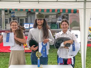 Two young woman and a young man standing behind a table, each holding a rabbit, papers, and blue ribbons, food truck in the background, trophy on the table