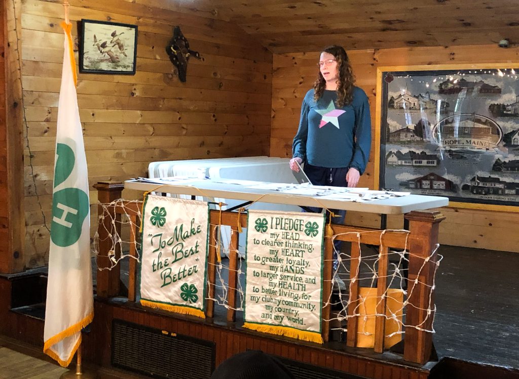 Young woman on a stage behind a table with 4-H flag and banners speaking