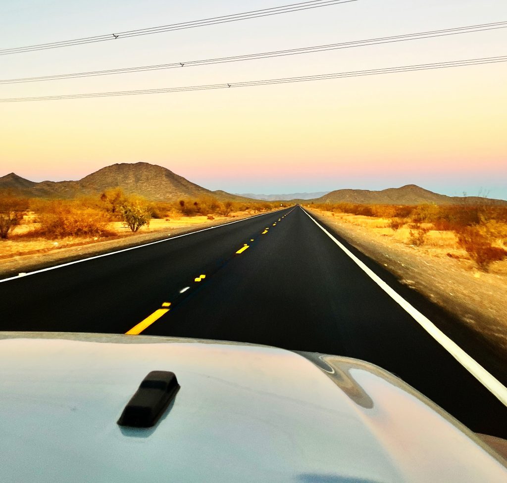 view of a black paved road in a dessert landscape with hills and sunset colors in the distant skyline, white vehicle hood in the foreground, powerlines above
