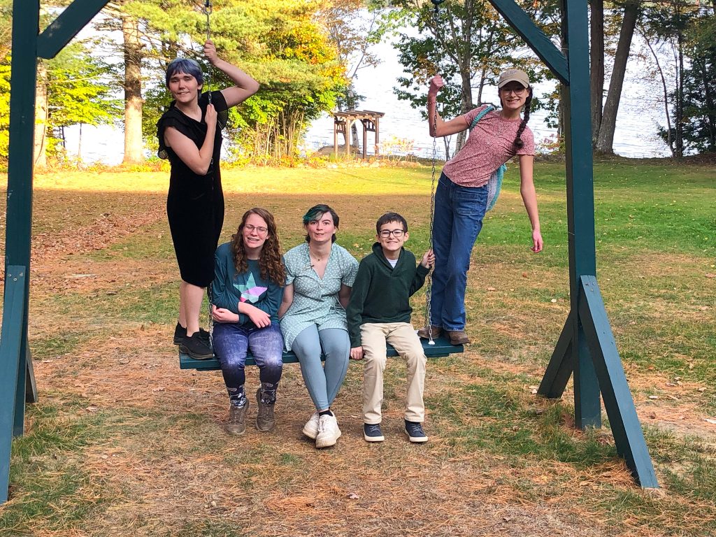 three young people sitting on a wooden swing, two young people standing on either side of swing in autumn landscape with lake in the distance