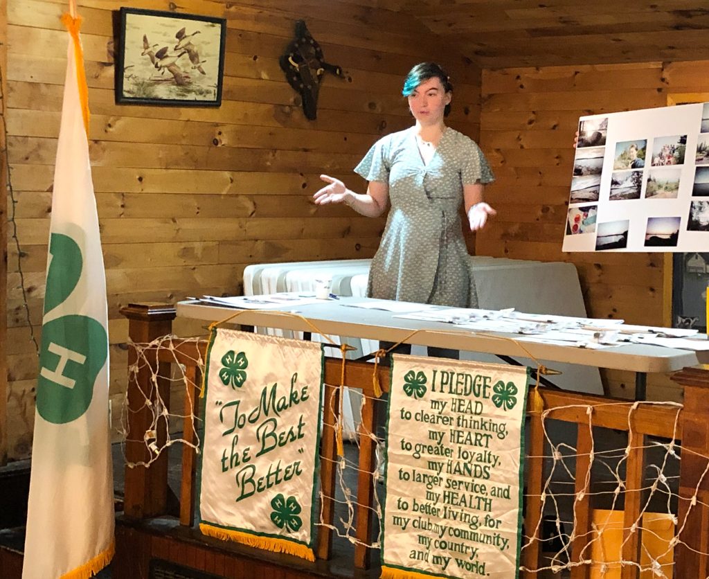 Young woman standing behind a table on a stage with a 4-H flag and banners, speaking