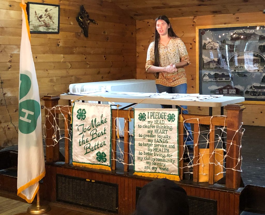Young woman on a stage behind a table with 4-H flag and banners speaking