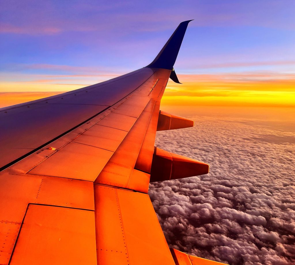view of an airplane wing from inside the plane, colored dramatically by the sunset colors also int he sky, gray clouds below