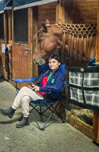 Young woman sitting in a chair in front of a barn stall containing a brown horse