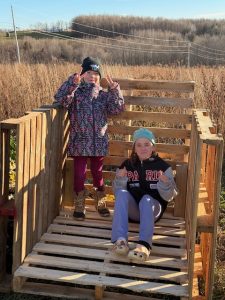 2 children, outdoors in a meadow, one standing and one sitting in an open-front structure built with wooden pallets