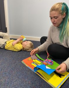 an adult reading a book to toddler laying on the floor of the Rumpus room