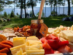cheese, fig jam in a jar and strawberries on a table by a lake that is surrounded by trees