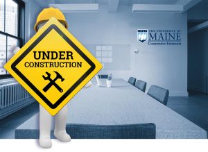 a cartoon figure holding an "under construction" sign in front of an empty conference room that is branded with UMaine Extension logo