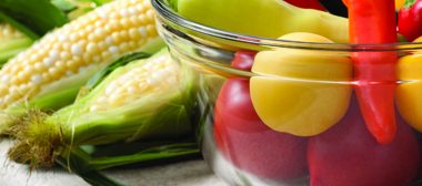 A photo of corn on the cob and a clear bowl full of fresh vegetables of various peppers
