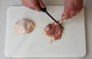 Photo of raw chicken showing removal of the bone from the thigh and how to use knife to scrape down the bone.