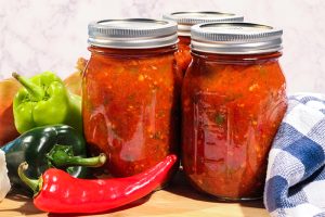 Three mason-type canning jars of tomato salsa on wood butcher block, a checked blue napkin, and several green and red peppers.