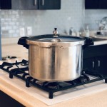 Silver pressure canner sitting on a stovetop in a kitchen with grey tile and brown cabinets