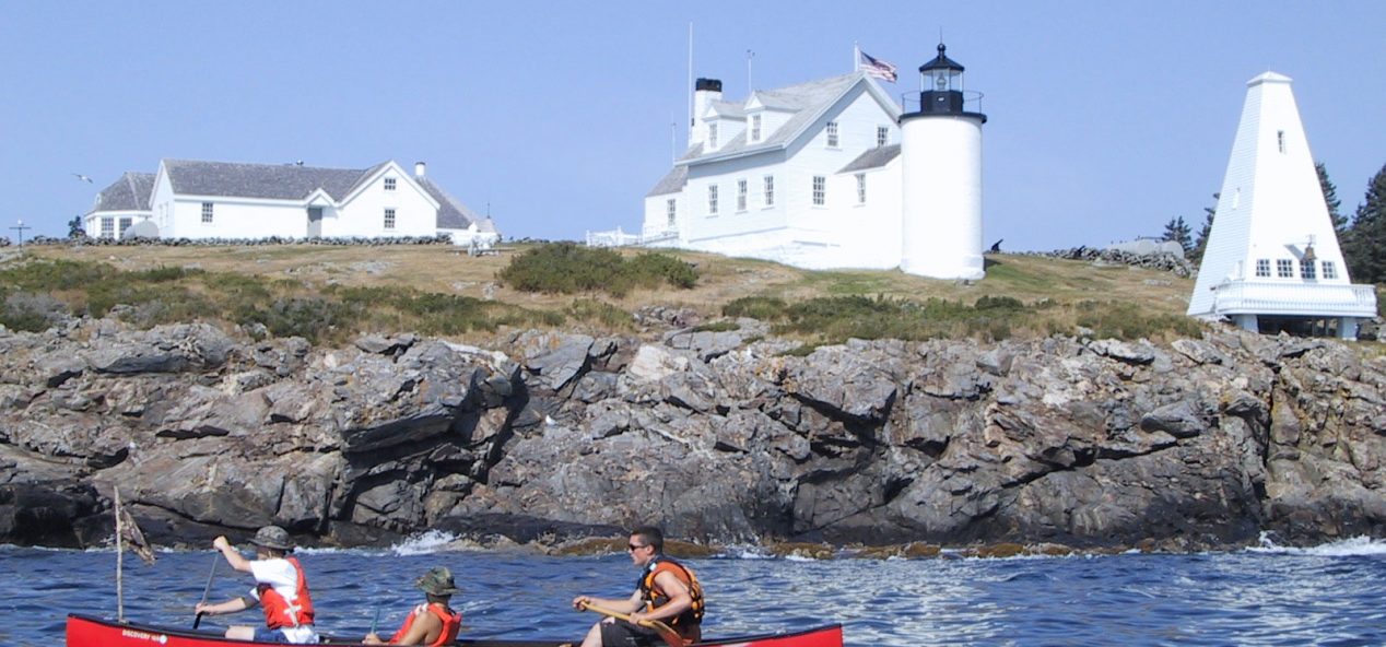 3 teens paddle canoe across the bay in front of lighthouse