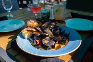 a plate of steamed mussels with garlic bread on the side and cocktails on the table in the background