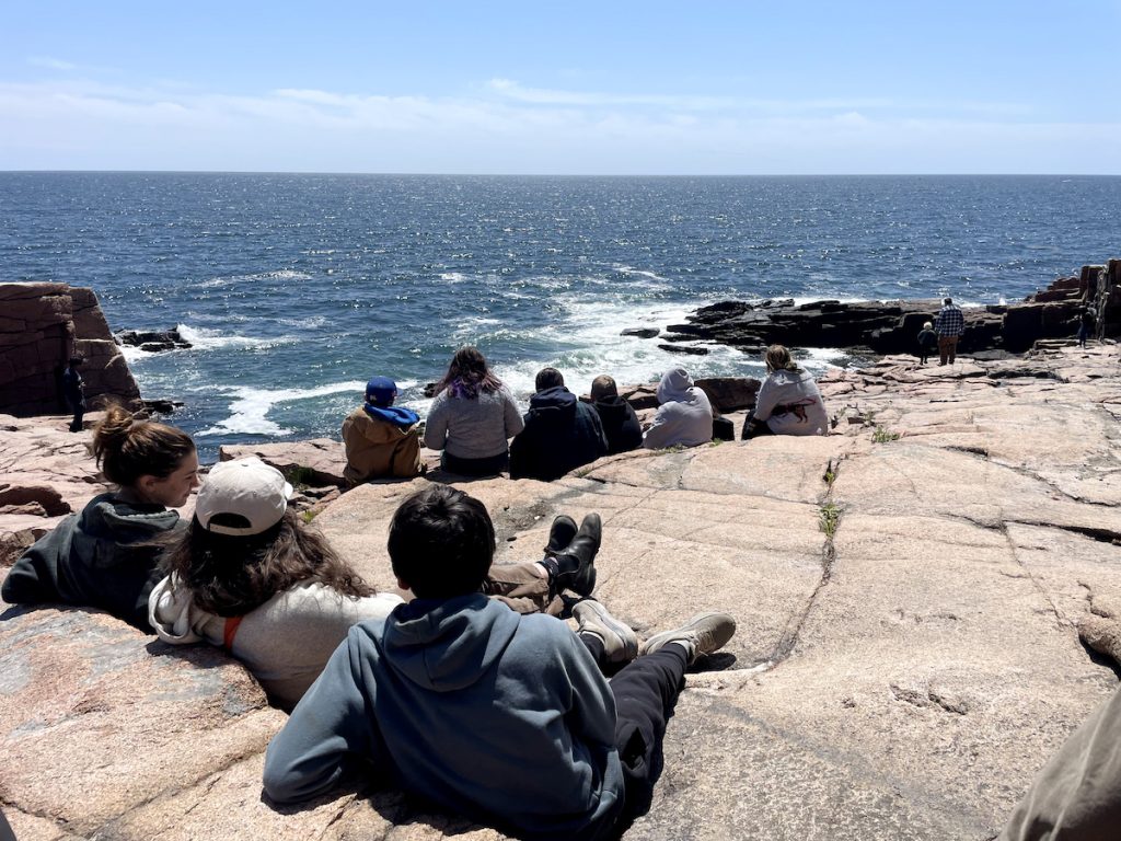 Group of teenagers sitting on a rocky coastline, looking out at the ocean.