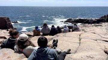 Group of teenagers sitting on a rocky coastline, looking out at the ocean.